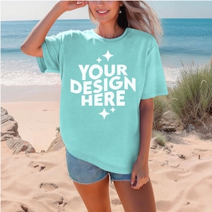 May include: A light turquoise t-shirt with the text "YOUR DESIGN HERE" in white. The shirt is worn with denim shorts on a sandy beach with the ocean in the background.