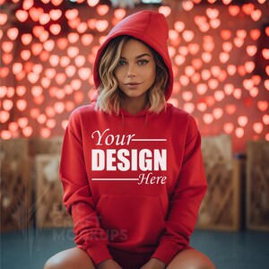 May include: A red hooded sweatshirt with the text "Your DESIGN Here" printed on the front. The model is wearing the sweatshirt and sitting in front of a background of red heart-shaped lights.