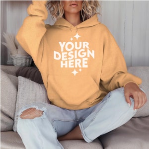 May include: A light yellow hoodie with a white print that says "YOUR DESIGN HERE" and stars. The hoodie is worn by a person sitting on a couch.