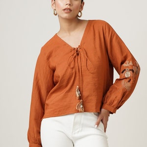 Puede incluir: Una mujer lleva una blusa de manga larga de color óxido con un lazo en el cuello y bordados en las mangas. También lleva unos vaqueros blancos acampanados y sandalias marrones de tacón.