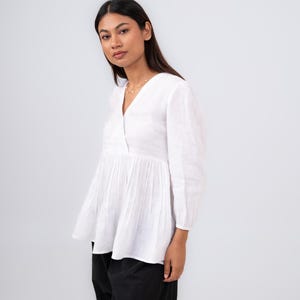 May include: A white linen top with a V-neck and three-quarter sleeves. The top features a gathered waist and a flowing, lightweight design. The model is wearing black pants. This top is suitable for casual or semi-formal wear.