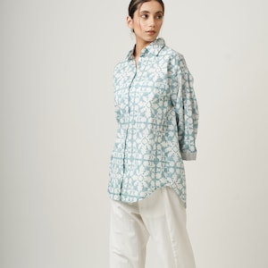 Puede incluir: Una camisa de manga larga azul claro con estampado blanco y cuello. La camisa tiene un corte holgado y está metida en un pantalón blanco.