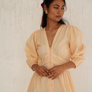 Puede incluir: Un vestido largo de manga larga de color melocotón con escote en V y cierre de botones en la parte delantera. El vestido tiene un corpiño ajustado y una falda fluida.