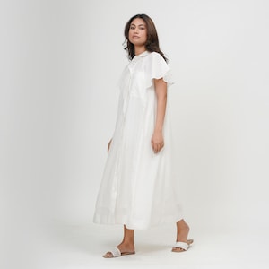 Puede incluir: Un vestido blanco de botones con mangas abullonadas. El vestido es de largo hasta la rodilla y tiene un corte holgado.