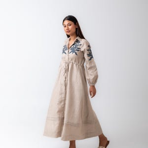 May include: A beige linen dress with long sleeves and a button-down front. The dress features a drawstring waist and is adorned with dark blue floral embroidery on the collar and sleeves. The model is wearing gold sandals.