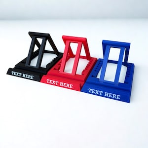 以下が含まれることがあります： 黒、赤、青の3色の携帯電話スタンド。各スタンドは、白い文字で「TEXT HERE」と印刷された長方形のベースを備えています。スタンドは、角度の付いたサポートと、電話やタブレットを保持するためのスロットで設計されています。