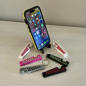 Puede incluir: Un teléfono inteligente con una funda amarilla descansa sobre un soporte negro. Varios llaveros abridor de botellas en varios colores, incluyendo rosa, gris, negro y blanco, están dispersos alrededor del teléfono. Los llaveros tienen un diseño de panal.