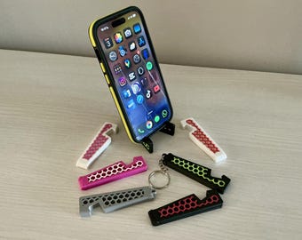 Può includere: Uno smartphone con una custodia gialla è appoggiato su un supporto nero. Diversi portachiavi apribottiglie in vari colori, tra cui rosa, grigio, nero e bianco, sono sparsi intorno al telefono. I portachiavi hanno un design a nido d'ape.