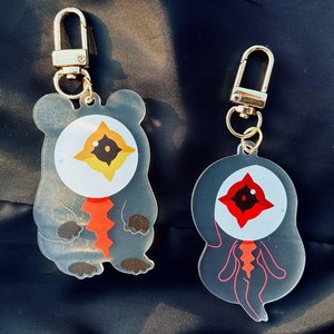 Puede incluir: Dos llaveros acrílicos con cierres dorados. Uno es una figura gris con forma de oso con un ojo grande y un diseño rojo y naranja. El otro es una figura gris con forma de fantasma con un ojo rojo y un diseño rojo y rosa.