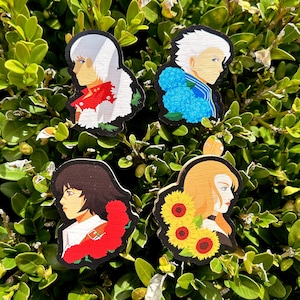 Puede incluir: Cuatro pines de madera con personajes y detalles florales. Los pines muestran un personaje de cabello blanco con flores rojas, un personaje rubio con girasoles, un personaje de cabello castaño con flores rojas y un personaje de cabello blanco con flores azules.