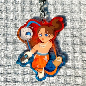 May include: A colorful acrylic keychain featuring a cartoon character with orange and blue clothing, holding a gun and a small robot. The character has brown hair and blue eyes. The keychain is attached to a silver metal ring.