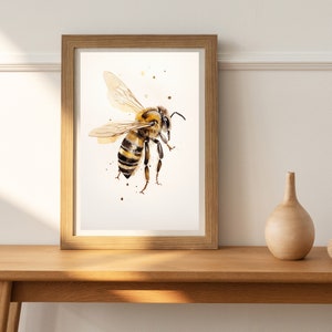 Op de afbeelding: Een aquarel schilderij van een bij met bruine en gele strepen, in een houten lijst. De bij is in de vlucht met zijn vleugels uitgespreid. De achtergrond is wit met bruine aquarel spatten.
