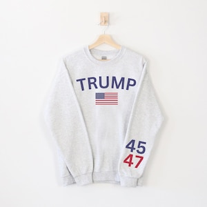 Pu&ograve; includere: Una felpa grigia con il testo "TRUMP" in lettere blu e una grafica della bandiera americana. I numeri "45" e "47" sono stampati sulle maniche in blu e rosso.
