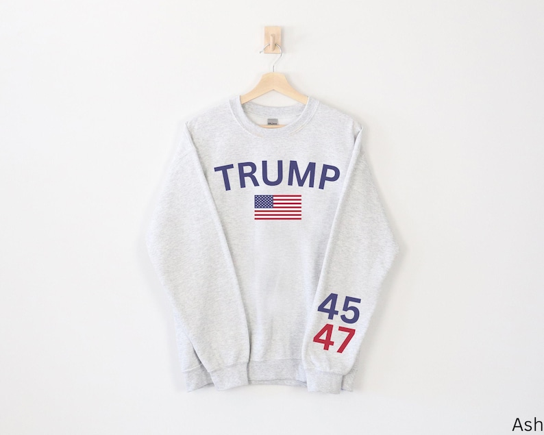 Pu&ograve; includere: Una felpa grigia con il testo "TRUMP" in lettere blu e una grafica della bandiera americana. I numeri "45" e "47" sono stampati sulle maniche in blu e rosso.