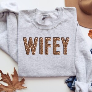 May include: A pale grey sweatshirt featuring the word "WIFEY" in brown, stitched lettering. A brown hat, a leaf, and denim shorts are visible in the background.