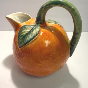 May include: A ceramic jug in the shape of an orange, featuring a green handle and leaves. The jug is orange with a textured finish, and the handle and leaves are green. The spout is white.