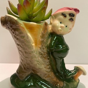 May include: A ceramic planter shaped like a tree stump with a green elf figurine holding a mushroom. The elf is wearing a red hat and green trousers. A succulent plant is growing in the planter.
