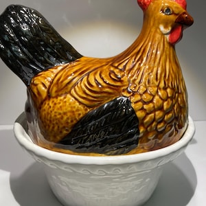 May include: A ceramic chicken figurine with brown and black markings, sitting in a white ceramic nest. The chicken has a red comb and a black eye.