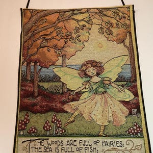 May include: A tapestry wall hanging featuring a fairy in a woodland scene. The fairy has wings and is wearing a yellow dress. The woodland is filled with trees, mushrooms, and a body of water. The tapestry includes the text "THE WOODS ARE FULL OF FAIRIES: THE SEA IS FULL OF FISH. THE TREES ARE FULL OF GOLDEN LEAVES: LET'S MAKE AN AUTUMN WISH, NANON."