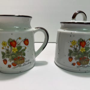 Peut inclure: Un ensemble de pot à crème et sucrier en céramique blanche. Chaque pièce présente un bord et une anse marron, avec un motif de fraises et de fleurs. Les mots "Wild Strawberry" sont imprimés.