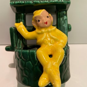 May include: A yellow ceramic elf figurine sitting in a green ceramic well planter. The elf has a red heart on its cheek and is wearing a yellow outfit.