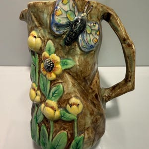 May include: A ceramic jug with a tree bark design, adorned with a butterfly and floral details. The butterfly has blue, yellow, and black markings. Yellow flowers and green leaves are also present. The handle is shaped like a tree branch.