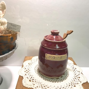 May include: A burgundy ceramic honey pot with a wooden spoon and a label that reads "Honey". The pot is sitting on a white crocheted doily.