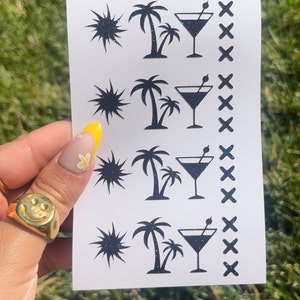 May include: A white rectangular sheet featuring black glittery illustrations of a sun, palm trees, martini glasses, and cross marks. The sheet is held against a green background.