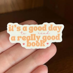 May include: A white sticker with orange text that reads "it's a good day to read a really good book" with two pink hearts on either side of the word "book".