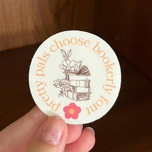 Puede incluir: Una pegatina redonda blanca con un contorno marrón y el texto "pretty pals choose bookerly font" en un círculo alrededor de una pila de libros con flores. Una flor rosa está en la parte inferior de la pegatina.