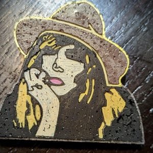 Puede incluir: Un posavasos de corcho con una ilustración en negro y amarillo de una mujer con un sombrero marrón. La mujer mira hacia abajo y tiene la mano en la barbilla.
