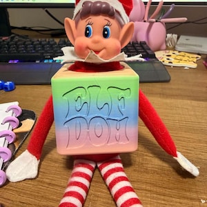May include: A plush elf doll with a red hat and red and white striped legs, wearing a square wooden block around its torso. The block features a gradient of green, blue, and pink with the embossed text ELF DOM on the front surface.