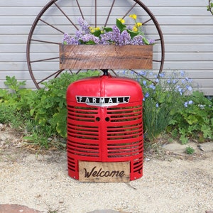May include: A red vintage tractor grill planter with a wooden box filled with purple and yellow flowers. The planter has a wooden sign that says "Welcome".