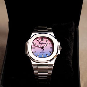 May include: A silver wristwatch with a pink and blue gradient dial. The watch face has the text "Fideliores Automatic" and a date window. The watch is in a black box.