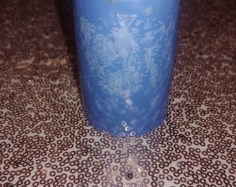 May include: A cylindrical blue pillar candle with a white top layer featuring embedded dried botanical pieces. The wax surface displays a mottled, crystalline texture throughout the body, and a single white cotton wick is centered at the top.