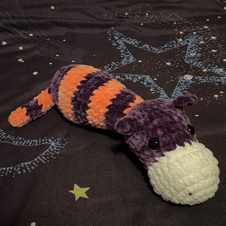 May include: A crocheted plush toy shaped like a long-nosed animal with purple and orange stripes. The toy has a white crocheted nose and black eyes.