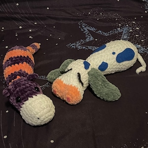 May include: Two crocheted stuffed animals, one a purple and orange striped worm and the other a white, blue, and green dog with a pink nose.