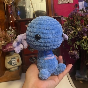 May include: A handmade blue crochet creature with black button eyes and lavender appendages. The plush toy has a round head and a small body with rounded feet. The creature is held in a hand.