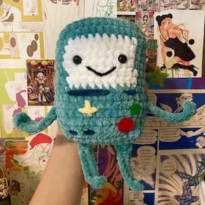 May include: A teal crochet plush toy shaped like a rectangular video game console. It features a white face, black button eyes, a black stitched smile, and colorful felt shapes including a yellow star, blue triangle, and red circle on the front.