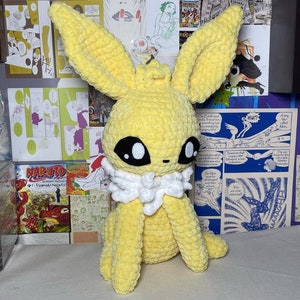 May include: A crocheted yellow Eevee plush toy with white fur around its neck. The toy has black eyes and a yellow body.