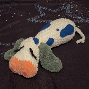 May include: A white crocheted stuffed animal with blue spots and gray ears. The animal is lying on its side with its eyes closed.
