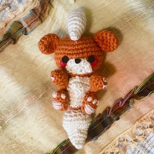 May include: A crocheted plush toy in the shape of a brown and white fox with a white tail. The toy has red eyes and a white nose.