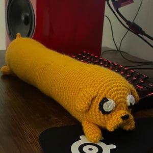 May include: A crocheted yellow dog-shaped pillow with white eyes and a black nose. The pillow is lying on a wooden surface with a black keyboard and a black mouse pad in the background.
