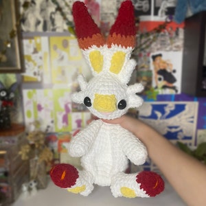 May include: A white crocheted plush toy with red and yellow accents. The toy has a long, pointed tail and large, round eyes. It is sitting on a person's hand.