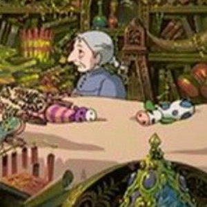 May include: A cluttered shop interior with a white table covered in a variety of colorful handmade toys. An older woman with gray hair sits at the table, looking to the left. The shop is filled with shelves of various items, including books, plants, and other crafts.