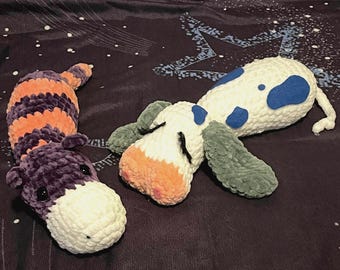 Puede incluir: Dos peluches hechos de hilo suave. Uno es una criatura a rayas moradas y naranjas con un hocico color crema y ojos negros. El segundo es una criatura blanca con manchas azules, orejas verdes y un hocico color melocotón con pestañas negras.