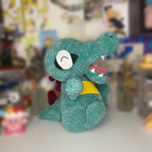 May include: A green crocheted plush toy shaped like a crocodile with a yellow chest and a pink tongue sticking out. The toy has a white eye with a black pupil and a black smile.