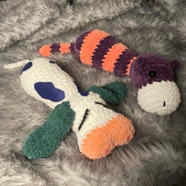 May include: Two crocheted stuffed animals, one is a purple and orange striped cat and the other is a white, green, blue and orange dog.