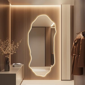 May include: A contemporary hallway with a large, organically shaped mirror with a glowing edge. The space features warm wood panelling, a neutral-toned cabinet with a vase of dried flowers, and a brown coat hanging on the wall. A light-coloured rug is on the floor.