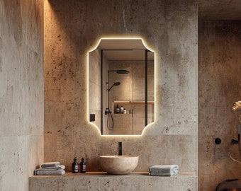 May include: A modern bathroom with a black and white vanity, a black toilet, and a large, rectangular mirror with a light-up border. The walls are a light beige color and the floor is a light brown color.